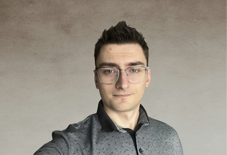 Portret mężczyzny w okularach, z ciemnymi, krótkimi włosami ułożonymi do góry, ubranego w szarą koszulę z kołnierzykiem, pełniącego funkcję Flutter Team Leader w itCraft.