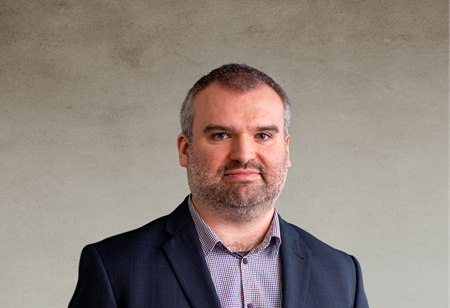 Portret mężczyzny z krótkimi, przerzedzonymi włosami i zarostem, ubranego w koszulę w kratę i ciemną marynarkę, pełniącego funkcję CEO w itCraft.