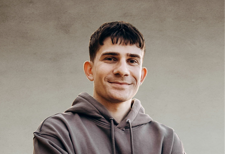 Portret uśmiechniętego mężczyzny z ciemnymi, krótkimi włosami z grzywką, ubranego w szarą bluzę z kapturem, pełniącego funkcję Frontend Developer w itCraft.
