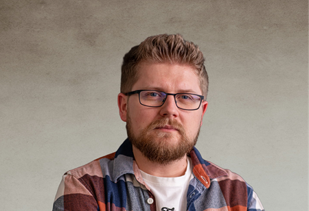 Portret mężczyzny w okularach, z krótkimi, zaczesanymi do góry jasnobrązowymi włosami i brodą, ubranego w biały t-shirt oraz kolorową koszulę w kratę, pełniącego funkcję Senior Android Developer w itCraft.