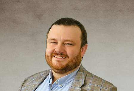 Portrait of a man with short dark hair and a beard, wearing a light shirt and a checked blazer, serving as Head of Discovery & Grow at itCraft.