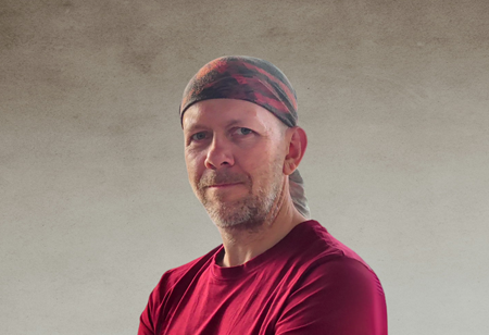 Portret mężczyzny z zarostem, ubranego w bordową koszulkę oraz kolorową bandanę na głowie, pełniącego funkcję iOS Tech Lead w itCraft.