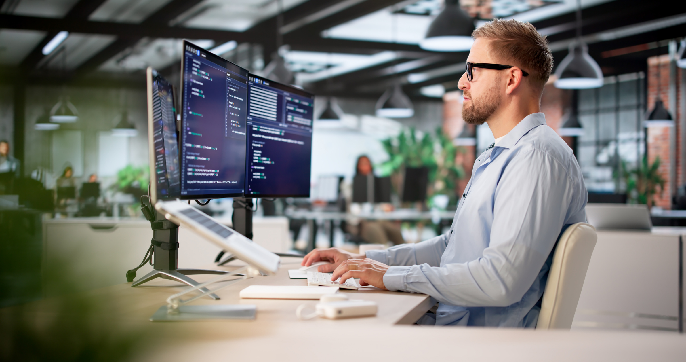 Mężczyzna w okularach pracujący w nowoczesnym biurze przed trzema monitorami wyświetlającymi linie kodu programistycznego.